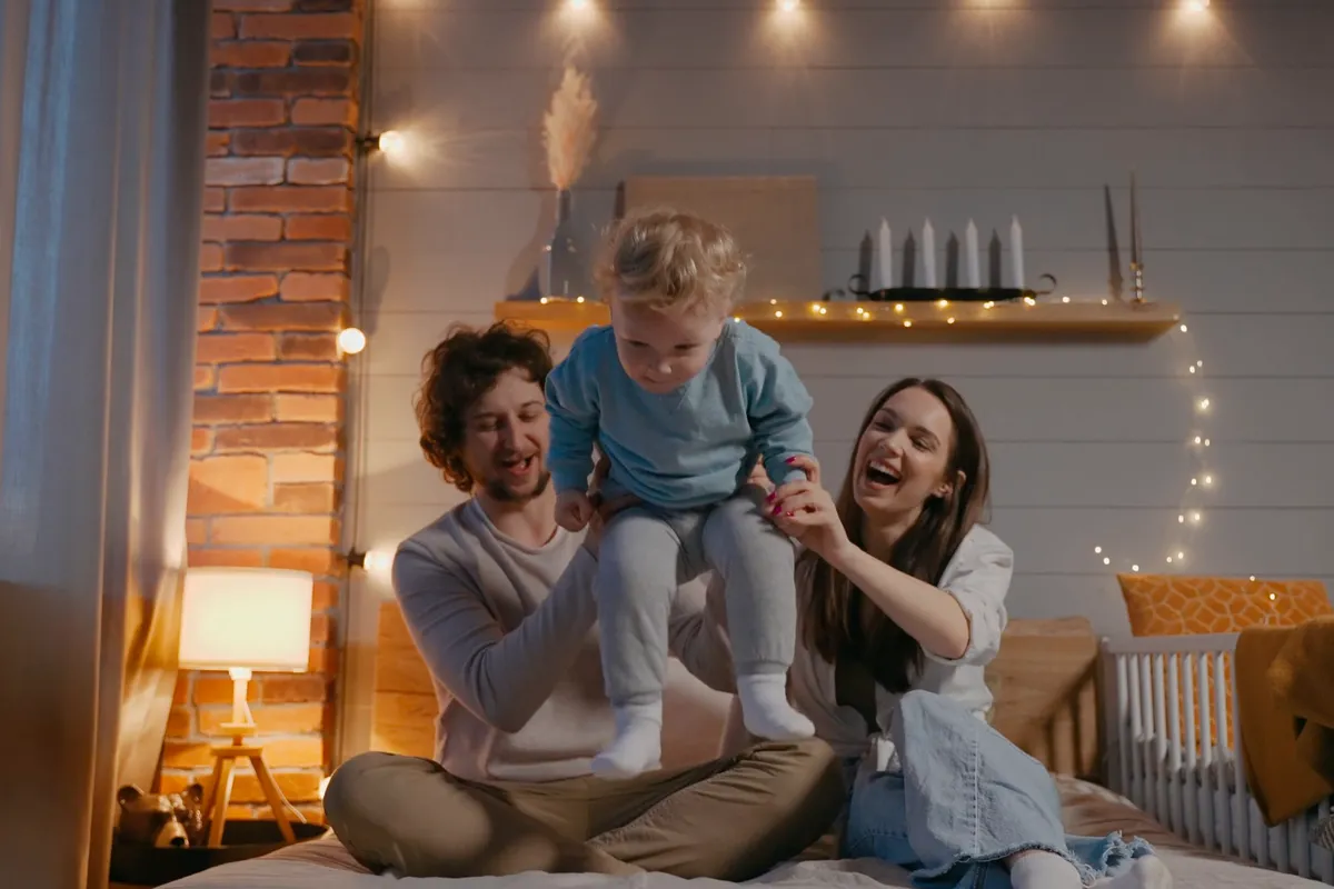 Parents laughing with their toddler on a bed in a warmly lit room